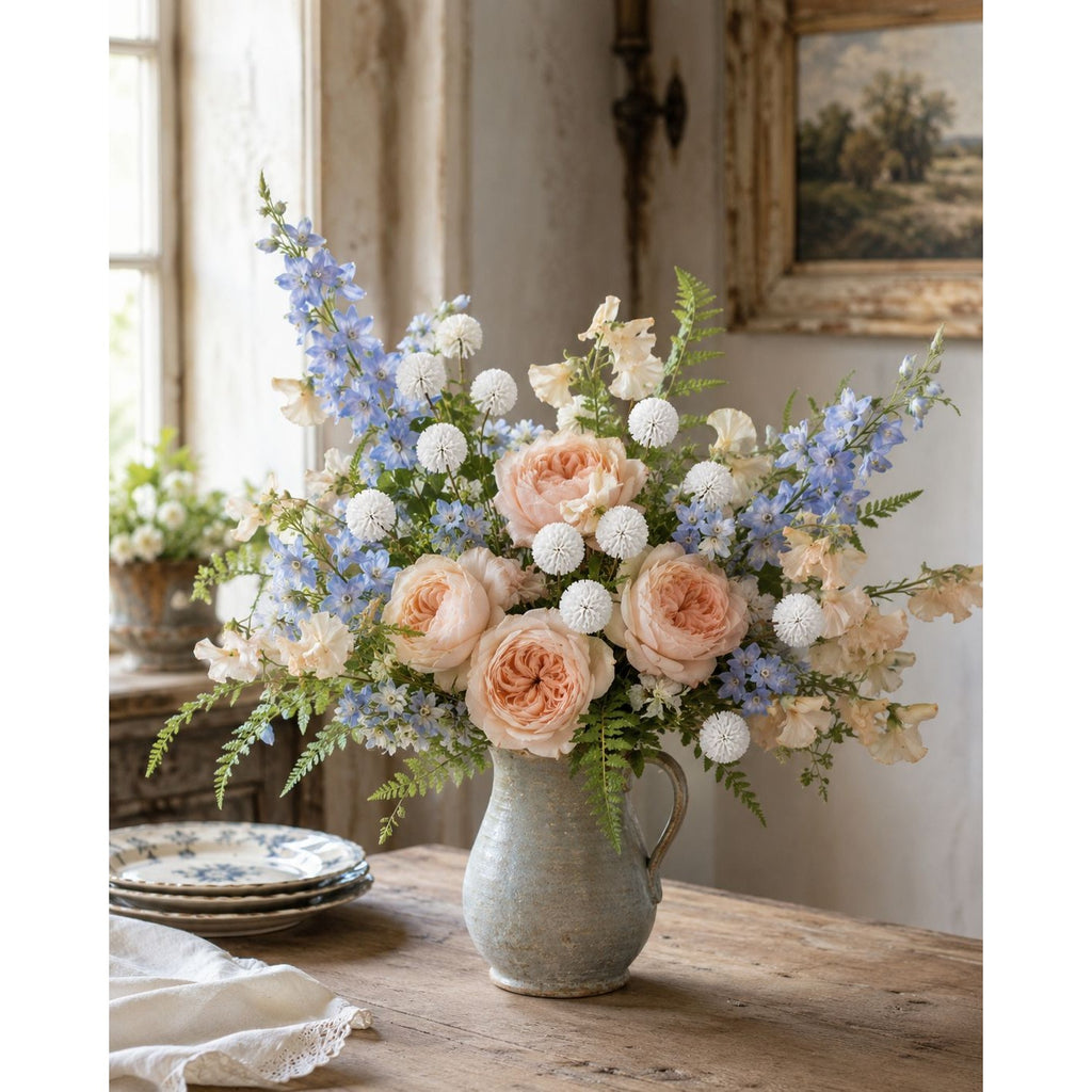 A floral arrangement in a rustic ceramic pitcher vase, featuring peach roses, blue delphiniums, and white pom-pom flowers, set on a wooden table in a softly lit room.