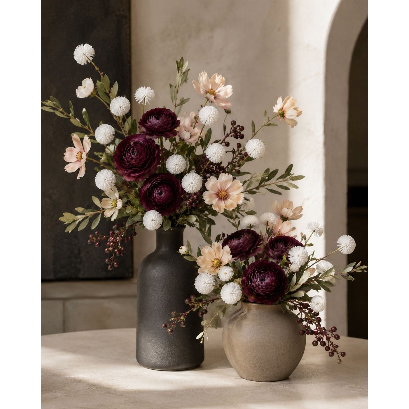 Two artificial flower arrangements in matte ceramic vases, displayed on a light stone surface in a softly lit interior setting. The arrangement features deep burgundy roses, blush peach daisies, white pom-pom flowers, and green foliage, with one larger dark charcoal vase and one smaller taupe-beige vase.