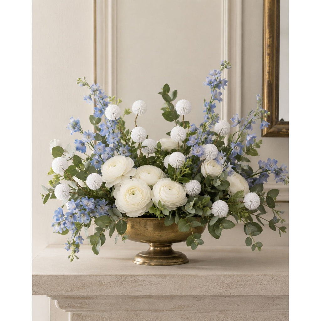A floral arrangement featuring white ranunculus, light blue delphiniums, and green eucalyptus in a gold pedestal bowl, displayed on a stone mantel against a beige paneled wall.
