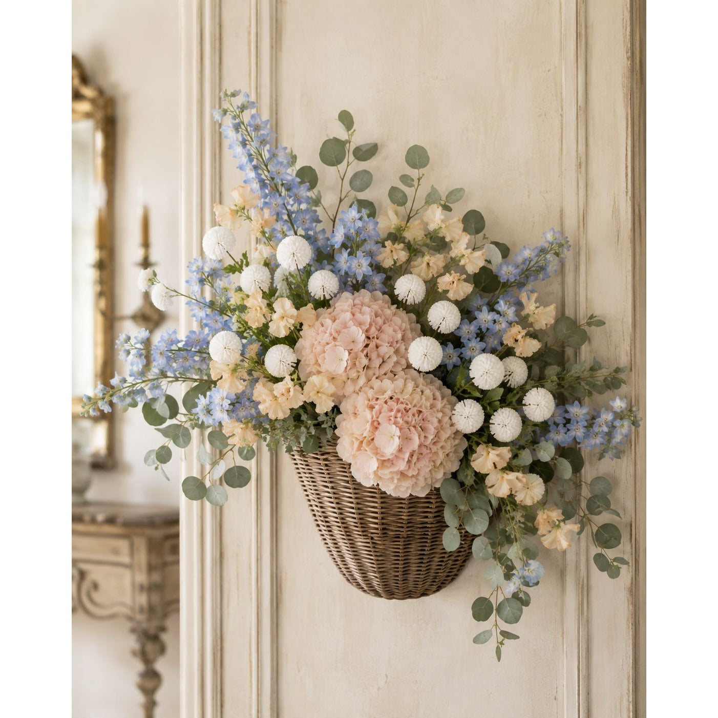 A wicker basket wall hanging filled with a floral arrangement of blush pink hydrangeas, light blue delphiniums, white pom-pom flowers, and green eucalyptus, displayed on a cream paneled wall.