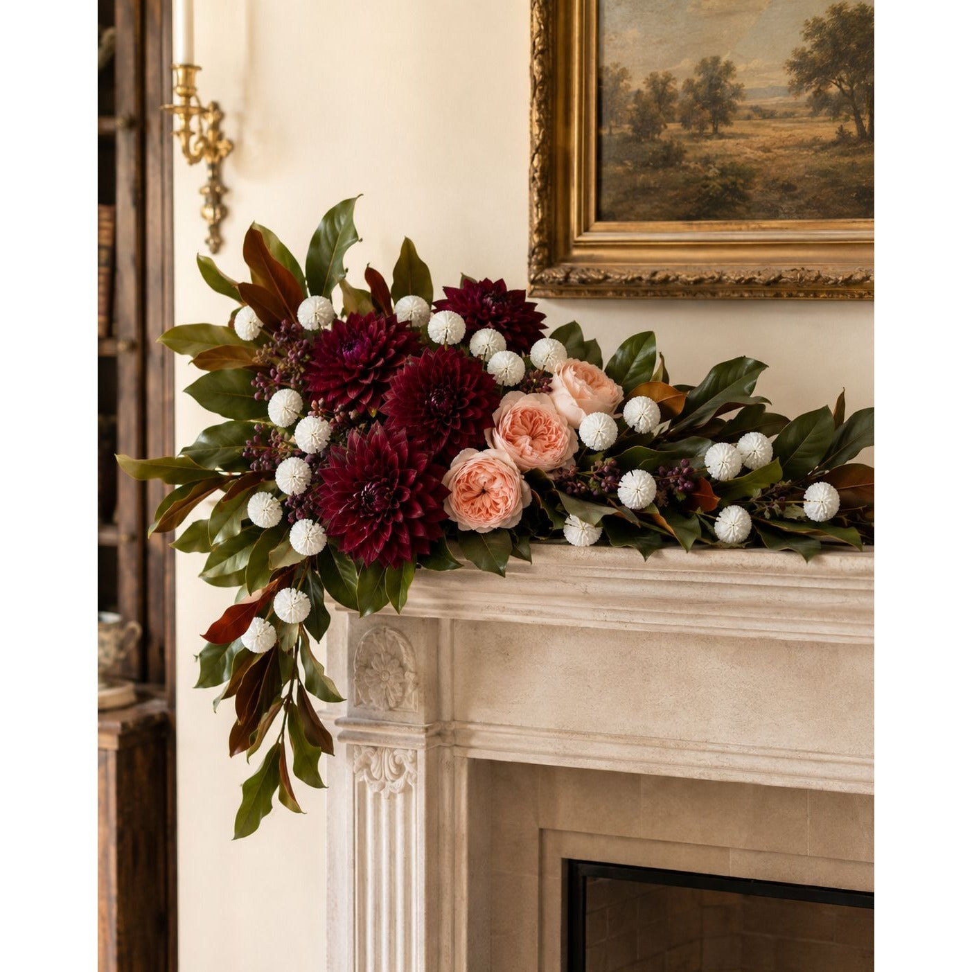 A large floral garland with deep burgundy dahlias, blush pink roses, white pom-pom balls, and green foliage draped over a cream-colored stone fireplace mantel in an elegant room.