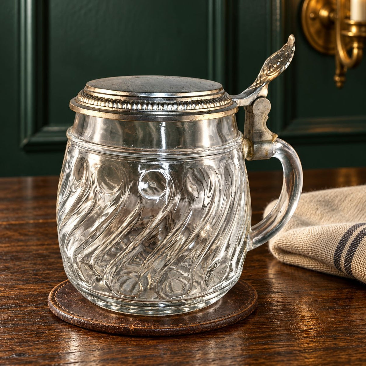 A clear glass beer mug with a silver pewter lid and handle, resting on a round wooden coaster on a dark wood table with a dark green paneled wall in the background.