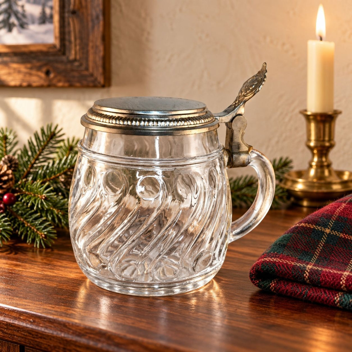 A clear glass beer mug with a silver pewter lid and handle, styled on a wooden table with a lit candle, pine branches, and a plaid cloth in the background.
