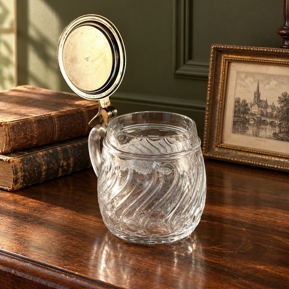 A clear glass mug with a handle and a hinged metal lid propped open, set on a dark wooden table beside old books and a framed picture in a room with green walls.