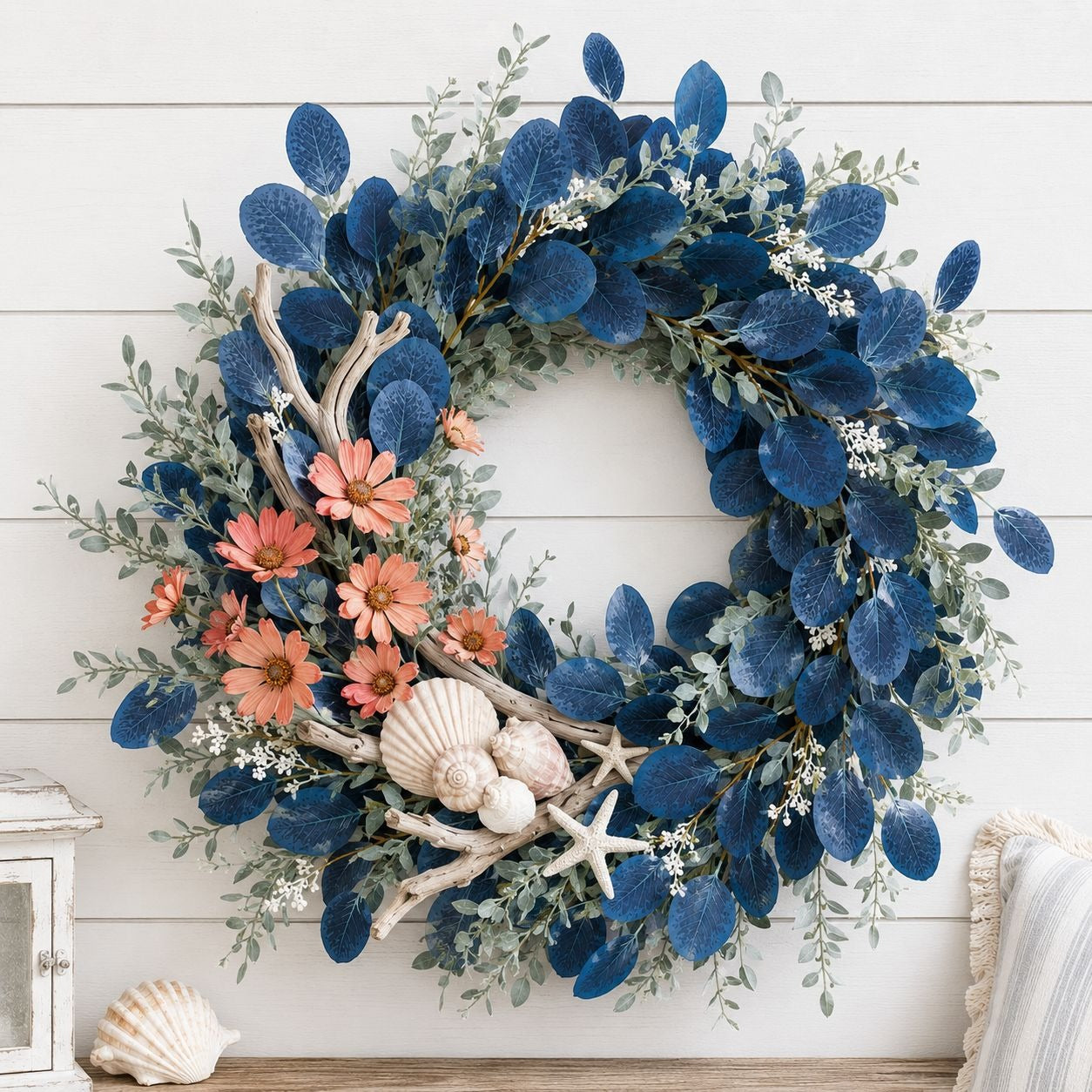 A decorative wreath with navy blue eucalyptus leaves, coral-orange flowers, white seashells, driftwood branches, and a starfish, hanging on a white shiplap wall.