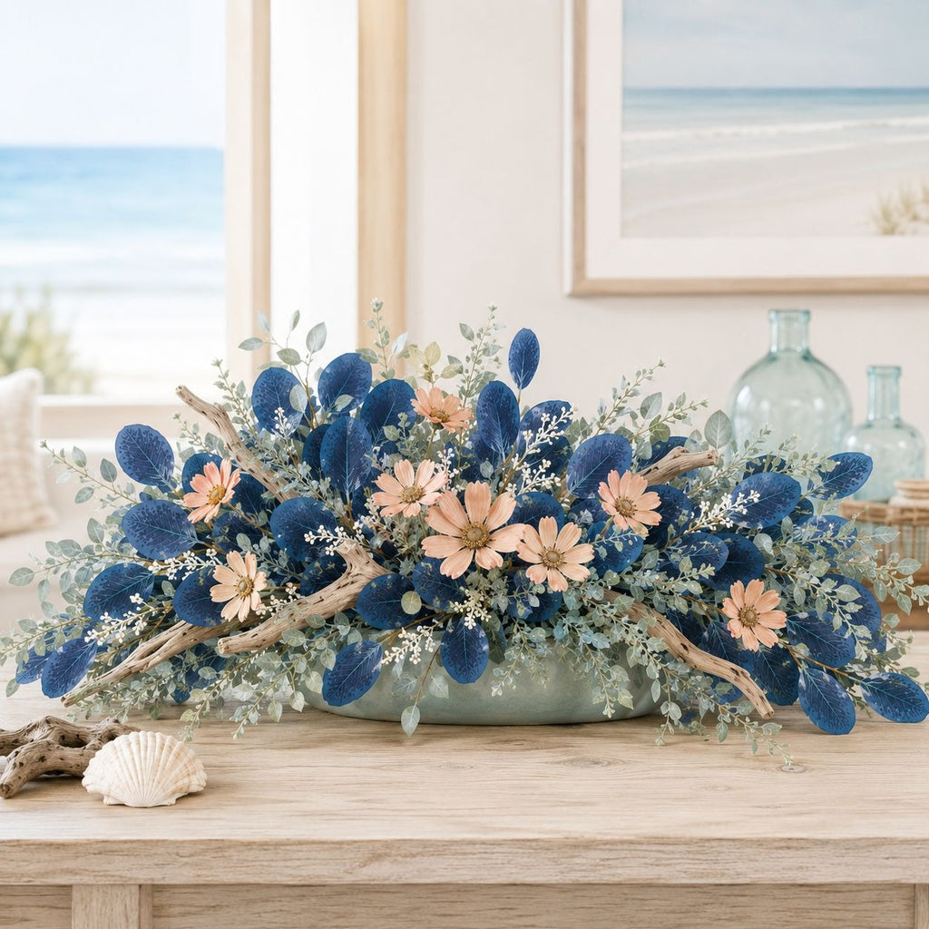 A coastal-themed floral centerpiece in a light blue-green ceramic bowl, featuring deep blue sea urchin clams, peach daisies, and sage green eucalyptus, styled on a wooden table with a seashell and glass bottles in a bright room.