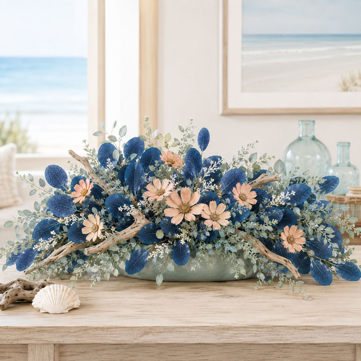 A coastal-themed floral centerpiece in a light blue-green ceramic bowl, featuring deep blue sea urchin clams, peach daisies, and sage green eucalyptus, styled on a wooden table with a seashell and glass bottles in a bright room.