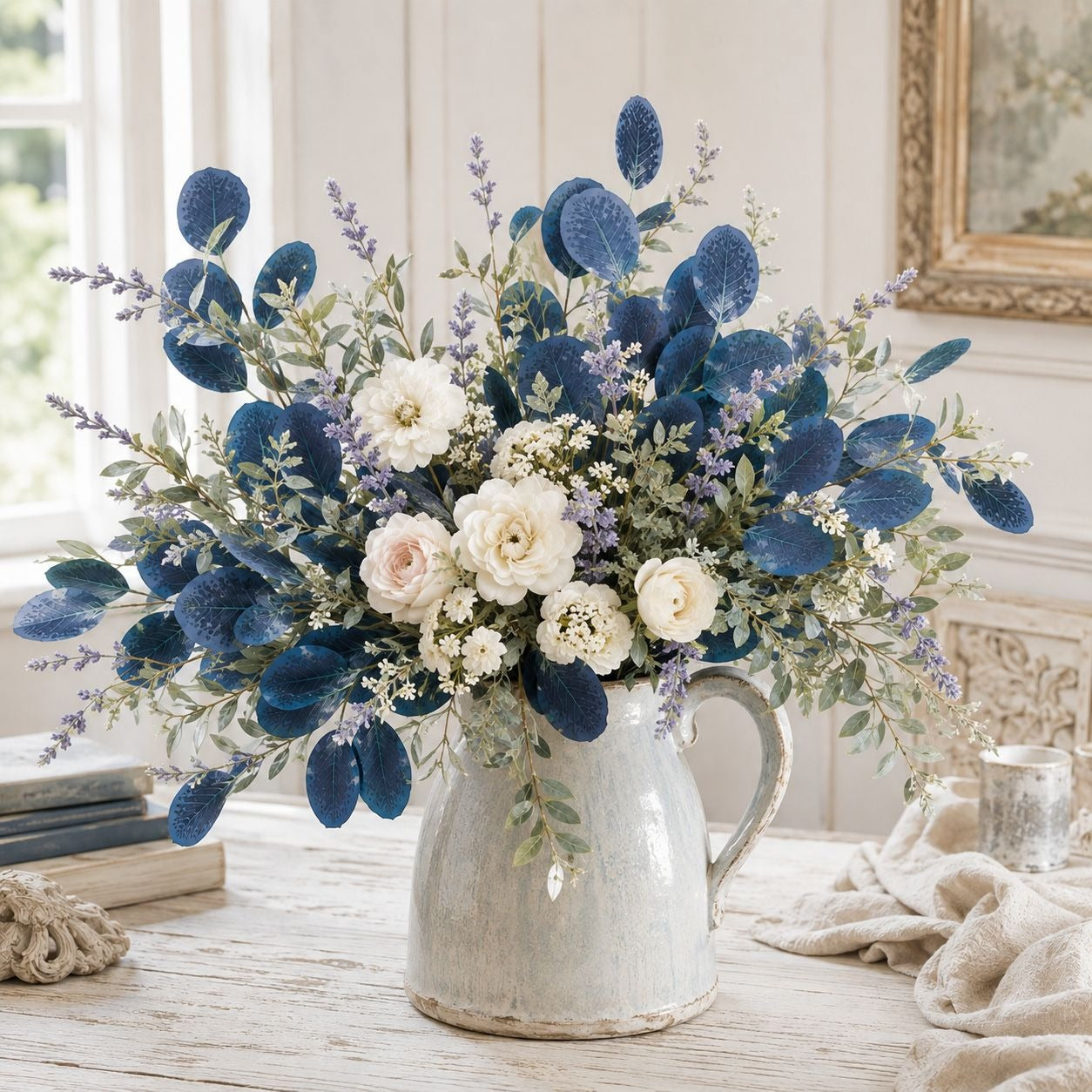 A large artificial floral arrangement in a distressed white ceramic pitcher vase, styled on a rustic wooden table with a neutral background.
