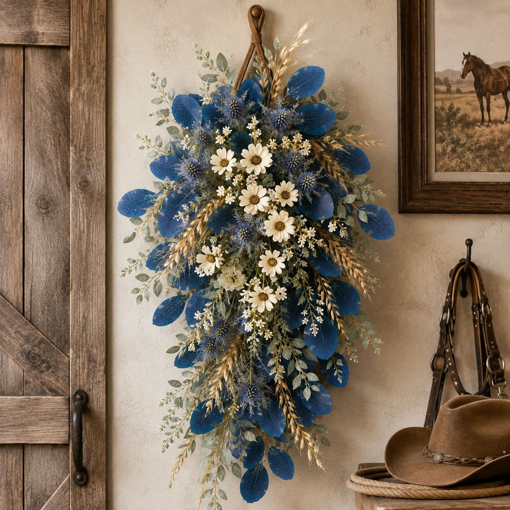 A rustic hanging floral arrangement with deep blue flowers, white daisies, and tan grasses, displayed on a beige wall beside a wooden door and framed western-style artwork.