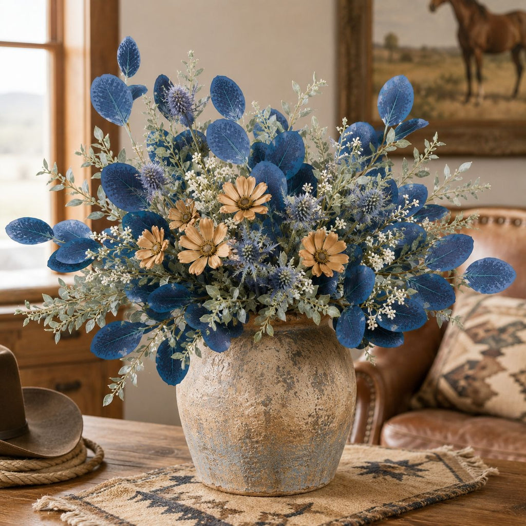 A rustic floral arrangement in a distressed ceramic vase, featuring prominent blue dried flowers, sage green foliage, and tan daisy-like blooms, displayed on a table in a warm, rustic living room setting.